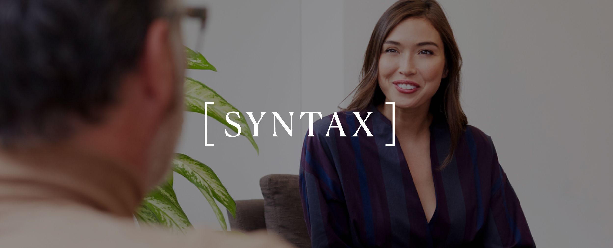 A woman and man are having a conversation in a professional setting. The Syntax logo is displayed over the image.