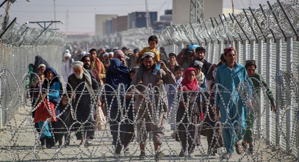 Photograph shows dozens of Afghan refugee families walking in a heavily fenced and barricaded area