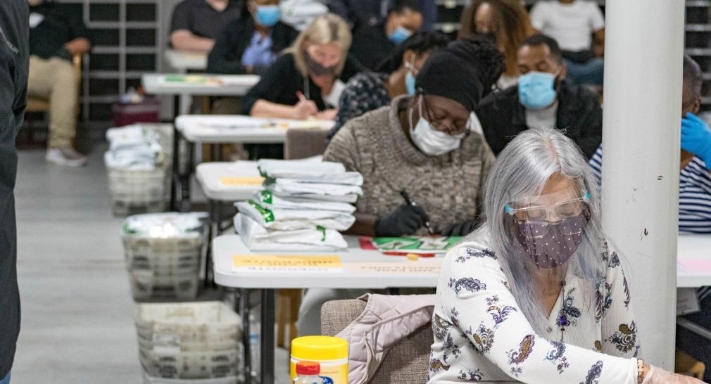 Photograph shows a room full of people recounting votes in Georgia. The photo features three workers in the foreground.