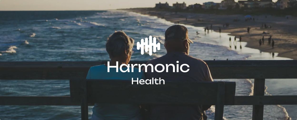 An older couple sits at a bench on a pier, looking out over a moderately active beach. The Harmonic Health logo appears over the image.